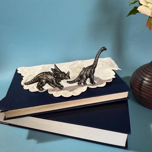 Puede incluir: Dos pequeñas y detalladas figuras de dinosaurios, un Triceratops y un Brontosaurus, descansan sobre un mantelito blanco encima de dos libros azul marino. Los dinosaurios son de color gris oscuro con superficies texturizadas. El fondo es azul claro.