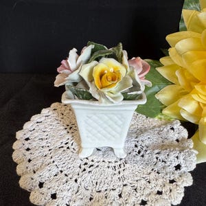 May include: A white ceramic planter with a lattice design, filled with porcelain roses in shades of pink, yellow, and white. The planter sits on a cream-coloured, crocheted doily. A large yellow flower is in the background.