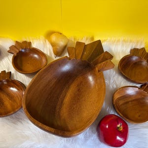 Puede incluir: Un cuenco de madera con forma de piña con seis cuencos más pequeños, también de madera, dispuestos sobre una superficie blanca y esponjosa. Manzanas rojas, una pera y una naranja están esparcidas alrededor de los cuencos sobre un fondo amarillo.