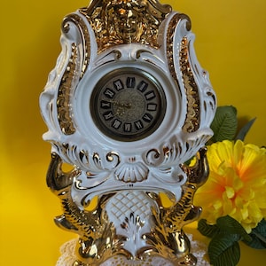 May include: A white and gold porcelain mantel clock with Roman numerals on the clock face. The clock has ornate gold detailing and a decorative face. The clock is sitting on a white lace doily.