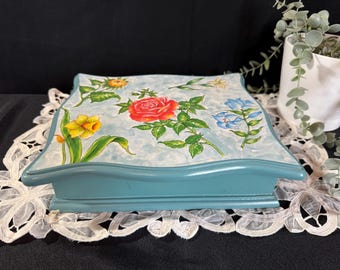 Puede incluir: Una caja de joyería cuadrada de madera con una base azul claro y un diseño floral en la tapa. La tapa presenta un fondo azul claro con flores pintadas, incluyendo una rosa roja, un narciso amarillo y flores azules.