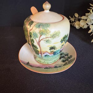 Puede incluir: Un azucarero de cerámica color crema con tapa y una pequeña cuchara de madera, colocado sobre un platillo a juego. El cuenco y el platillo están decorados con una escena de paisaje con árboles, una casa y un campo rosa y verde.