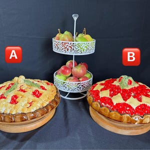 Puede incluir: Dos fuentes decorativas para tartas con tapas, una con corteza de rejilla y relleno rojo, la otra con diseño de fruta roja. Un soporte blanco de dos niveles contiene manzanas y peras. Los platos se asientan sobre bases de madera.
