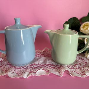 Puede incluir: Dos teteras de cerámica, una azul claro y otra verde menta, cada una con tapa y asa. Se encuentran sobre un tapete de encaje blanco sobre un fondo rosa. Rosas blancas y vegetación en el fondo.