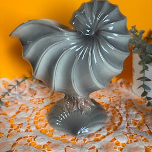 May include: A grey ceramic nautilus shell sculpture with a glossy finish. The sculpture has a spiral design and sits on a pedestal base. The background is a bright orange colour.