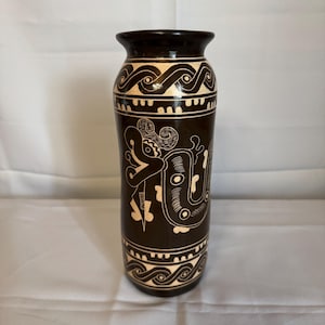 Puede incluir: Un jarrón alto de cerámica marrón oscuro con intrincados diseños blancos grabados. El jarrón presenta patrones repetidos de ondas y geométricos, junto con una figura central estilizada. El borde y la base también son de color marrón oscuro.