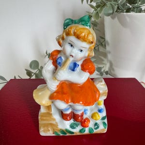 以下が含まれることがあります： フルートを演奏する少女のヴィンテージ磁器フィギュア。 少女はオレンジ色の髪、緑のリボンをしており、青いアクセントのオレンジ色のドレスを着ています。 フィギュアは、カラフルな花のディテールが施された木製のベンチに座っています。