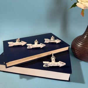 May include: White arrow-shaped cabinet knobs with a distressed finish are mounted on a dark blue book. The knobs have a silver stem. A vase with a white rose is in the background. The knobs are decorative.