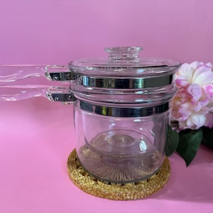 May include: A clear glass double boiler with a lid and two handles. The pot has a silver band around the middle and sits on a woven coaster. A pink and white flower is in the background.