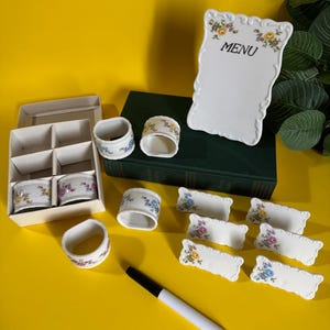 May include: A collection of white ceramic tableware with floral designs, including napkin rings, place card holders, and a menu board. The menu board has the word "MENU" printed on it. A black marker is also present.