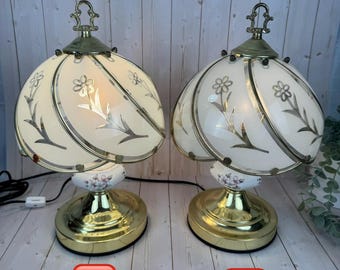 May include: Two vintage table lamps with a floral design. The lamps have a cream-colored shade with gold accents and a gold base. The shades are adorned with a floral pattern. The base has a floral design. The lamps are labeled A and B.