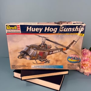 Könnte beinhalten: Modellbausatz-Box für einen Huey Hog Sunship Hubschrauber. Die Box zeigt eine detaillierte Illustration des Hubschraubers im Flug, mit dem Wort "MARINES". Das Set enthält ein Modell, Pinsel, Farben und Kleber.