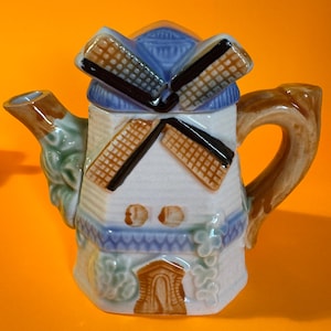 Può includere: Teiera in ceramica a forma di mulino a vento. Ha un corpo bianco con dettagli blu e marroni, un manico e un beccuccio marroni e pale del mulino marroni e nere. Il design include un tetto blu ed elementi decorativi.