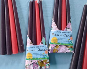 May include: Advent candles in a variety of colors, including red and dark purple. The candles are packaged in floral-patterned paper with the text "pure beeswax Advent Candle". The candles are approximately 10 inches tall.