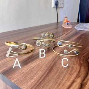 May include: Three gold-toned arm cuffs with gemstone accents. Cuffs feature unique designs, including spiral and teardrop shapes. Each cuff is labeled with a letter: A, B, and C. The jewelry is displayed on a dark wooden surface.