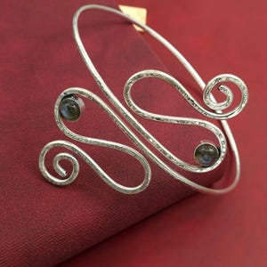 May include: Silver-toned arm cuff with a hammered texture and two small, round, iridescent gemstones. The cuff features a decorative, openwork design with swirling patterns. The jewelry is displayed on a red surface.
