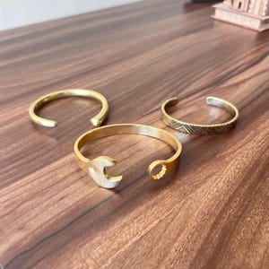 May include: Three gold-colored metal bracelets on a wooden surface. One is a simple open cuff, another features a wrench design, and the third has a geometric pattern. The bracelets are open-ended, allowing for easy wear.
