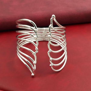 May include: A silver arm cuff with a distinctive design. It features multiple curved, parallel bands, creating a layered effect. The design includes a central connecting piece and flared ends. The cuff is set against a red background.