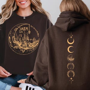 a woman wearing a black sweatshirt with a gold design on the front, which includes a moon and stars, and a back design with a series of crescent moons and stars.