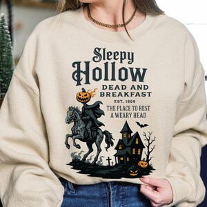 A woman wearing a beige sweatshirt with a Halloween-themed design featuring a skeleton on horseback, a castle, and jack-o-lanterns.