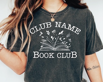 May include: Dark gray t-shirt with the words "CLUB NAME BOOK CLUB" in white, above an open book graphic. The design includes illustrations of leaves, dragonflies, and a small dragon. The shirt is a comfortable, casual style.