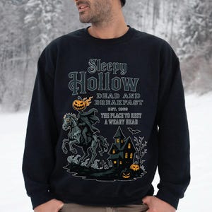 A man wearing a black sweatshirt with a Halloween-themed design, including a skeleton riding a horse, a castle, and jack-o-lanterns.