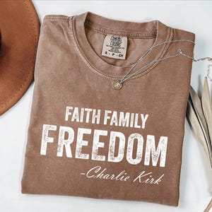 A brown t-shirt with the text "Faith Family Freedom" printed on it, along with a necklace and a hat.