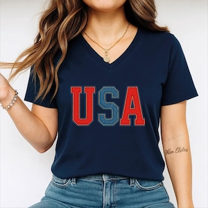 May include: Navy blue V-neck t-shirt with the word "USA" in a red and blue denim-style design. The shirt is a short-sleeved top, suitable for casual wear. The image shows a close-up of the shirt.