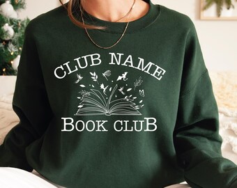 May include: Dark green sweatshirt with white text that reads "CLUB NAME BOOK CLUB". The design features an open book with illustrations of leaves, insects, and other small objects. The sweatshirt is a solid color.