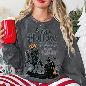A woman wearing a gray sweatshirt with a Halloween-themed design, including a pirate, a castle, and a jack-o-lantern. She is holding a red mug and appears to be sitting on a couch.
