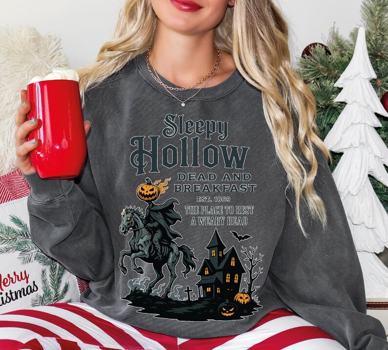 A woman wearing a gray sweatshirt with a Halloween-themed design, including a pirate, a castle, and a jack-o-lantern. She is holding a red mug and appears to be sitting on a couch.