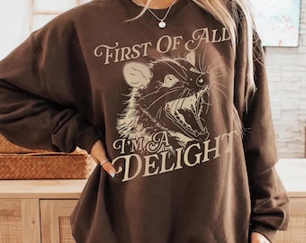 May include: A brown sweatshirt featuring a detailed illustration of a possum with the text "First Of All I'm A Delight" in a vintage font. The design is in a cream color, contrasting with the dark brown fabric.