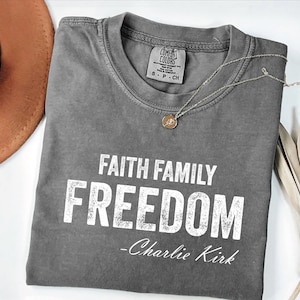 A gray t-shirt with the text "FAITH FAMILY FREEDOM - Charlie Kirk" printed on it, along with a necklace and a brown hat.