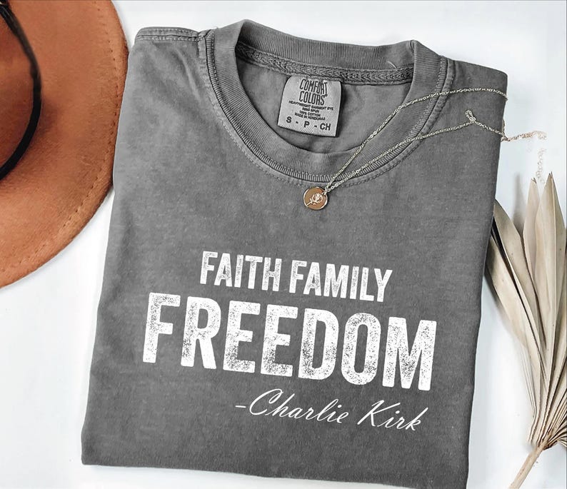 A gray t-shirt with the text "FAITH FAMILY FREEDOM - Charlie Kirk" printed on it, along with a necklace and a brown hat.