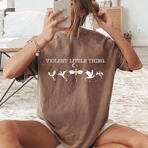 A person is sitting on the floor, holding a smartphone up to their face, taking a selfie. They are wearing a brown t-shirt with a graphic design and text printed on it.