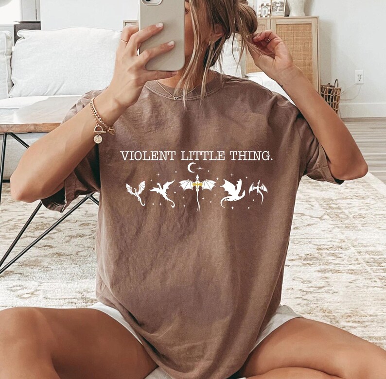 A person is sitting on the floor, holding a smartphone up to their face, taking a selfie. They are wearing a brown t-shirt with a graphic design and text printed on it.