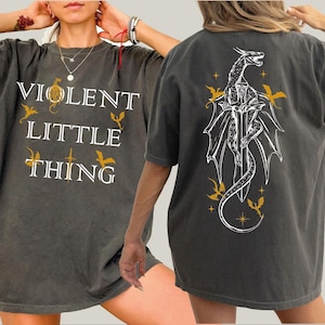 May include: Dark gray t-shirt with the text "VIOLENT LITTLE THING" in white and gold. The back features a white dragon design with gold accents. A casual, oversized tee.