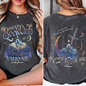 May include: A charcoal gray t-shirt featuring a front graphic with a mountain, stars, and the text "House of Wind Book Club Est. 21 Valkyrie reading room." The back graphic shows a sword, crescent moon, and "I am the rock against which the surf crashes. Nothing can Break me."