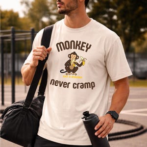 May include: Cream-colored t-shirt with a cartoon monkey eating a banana. The shirt has the text "MONKEY" above the monkey graphic and "never cramp" below. The shirt is worn by a person carrying a black gym bag and water bottle.