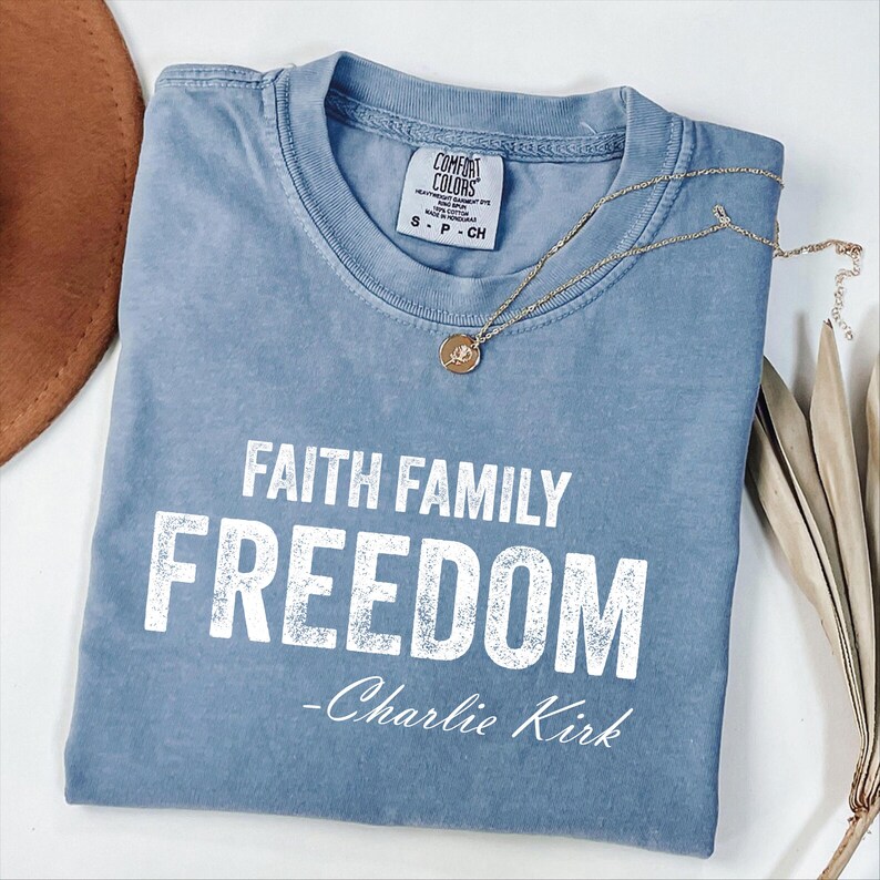 A blue t-shirt with the text "FAITH FAMILY FREEDOM - Charlie Kirk" printed on it, along with a necklace and some leaves.