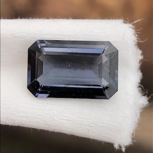 May include: A rectangular, faceted gemstone with a deep blue-gray hue. The gem is set against a white, textured background, showcasing its sharp edges and polished surface. The cut of the stone is an emerald cut.