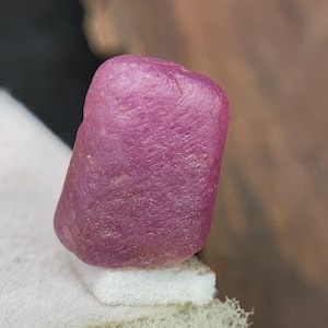 Puede incluir: Primer plano de una gema de rubí en bruto sin cortar. El rubí es de color rojo rosado intenso con una superficie texturizada. La gema descansa sobre una superficie blanca, con un fondo borroso marrón.