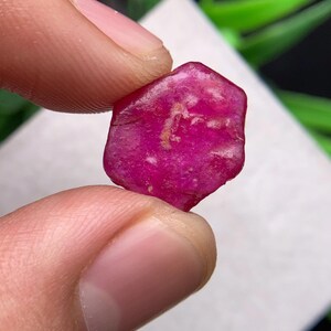 Può includere: Una gemma di rubino grezza di forma esagonale con una vibrante tonalità rosso-rosa intenso. La superficie della pietra presenta una consistenza naturale non lucidata con sottili variazioni di colore e piccole inclusioni chiare.