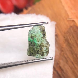 Könnte beinhalten: Ein roh geschliffener Smaragd, gehalten von einer Pinzette. Der Smaragd zeigt einen leuchtenden Grünton mit dunkleren, strukturierten Bereichen. Der Edelstein liegt auf einer weißen Oberfläche mit unscharfem Hintergrund.
