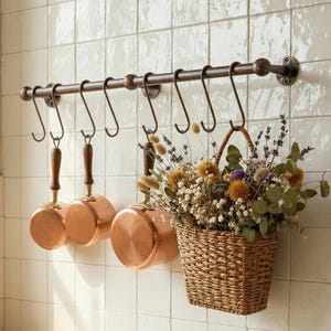 May include: A kitchen scene features a bronze-colored pot rack with hanging copper pots and a woven basket filled with dried flowers. The pots have wooden handles. The rack is mounted on a white tiled wall, creating a rustic, farmhouse aesthetic.