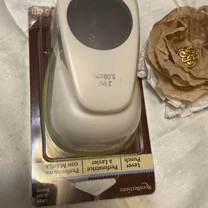 May include: A white and green lever punch by Recollections, measuring 5.08 cm. The punch has a circular cutting area and is packaged in a black and brown cardboard backing. A decorative paper flower with a gold embellishment is also visible.