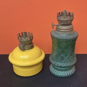 Può includere: Una base per lampada a olio gialla con finitura liscia e una lampada a olio in vetro verde con motivo a rombi testurizzato e base smerlata. Entrambe le lampade presentano componenti del bruciatore in metallo con una parte superiore a corona.