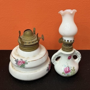 Puede incluir: Una base de lámpara de aceite de cerámica blanca grande con quemador de latón y patrones de rosas rosas, junto a una lámpara de aceite de cerámica blanca más pequeña con dos asas, diseño de rosas y pantalla de cristal blanco con volantes.
