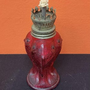 Könnte beinhalten: Eine rote Öllampe aus Glas in Fischform mit texturiertem Schuppenmuster und einem runden Sockel. Oben befindet sich ein metallener Brenner mit gezacktem Rand und einem weißen Stoffdocht, der aus der Mitte des Metallkragens herausragt.