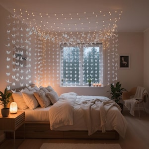 Puede incluir: Un dormitorio acogedor con una cama, almohadas y una manta suave. Cadenas de luces de hadas y grullas de papel cuelgan del techo y frente a la ventana, creando un ambiente cálido. Una pequeña mesita de noche de madera con una lámpara está junto a la cama.